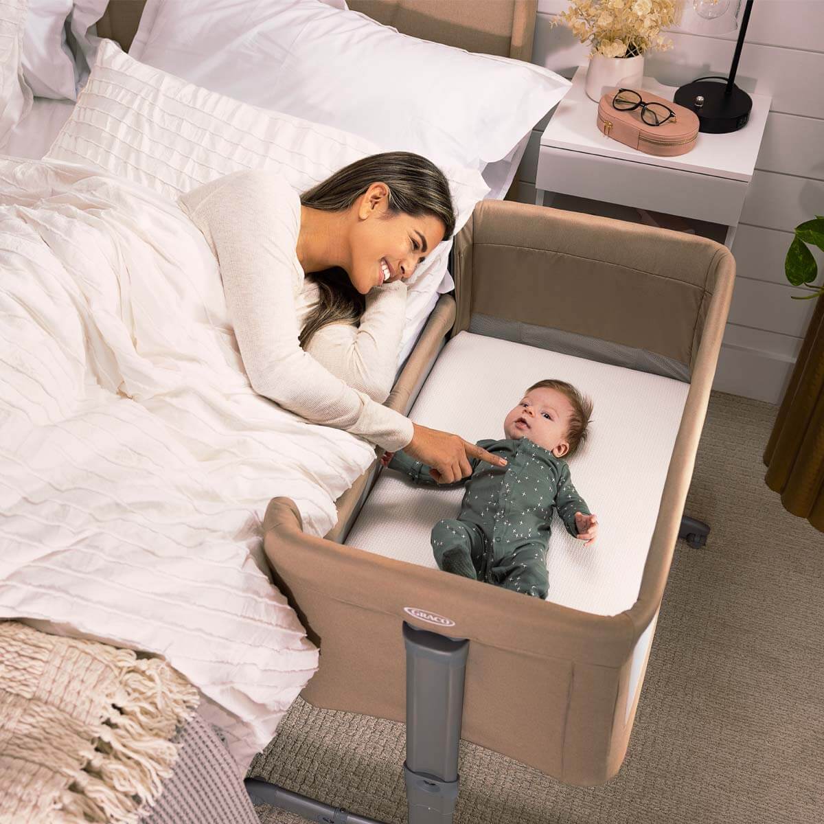 Mama sieht aus dem Elternbett heraus nach dem Baby, dass im Graco® Sweet2Sleep™ Beistellbett liegt.