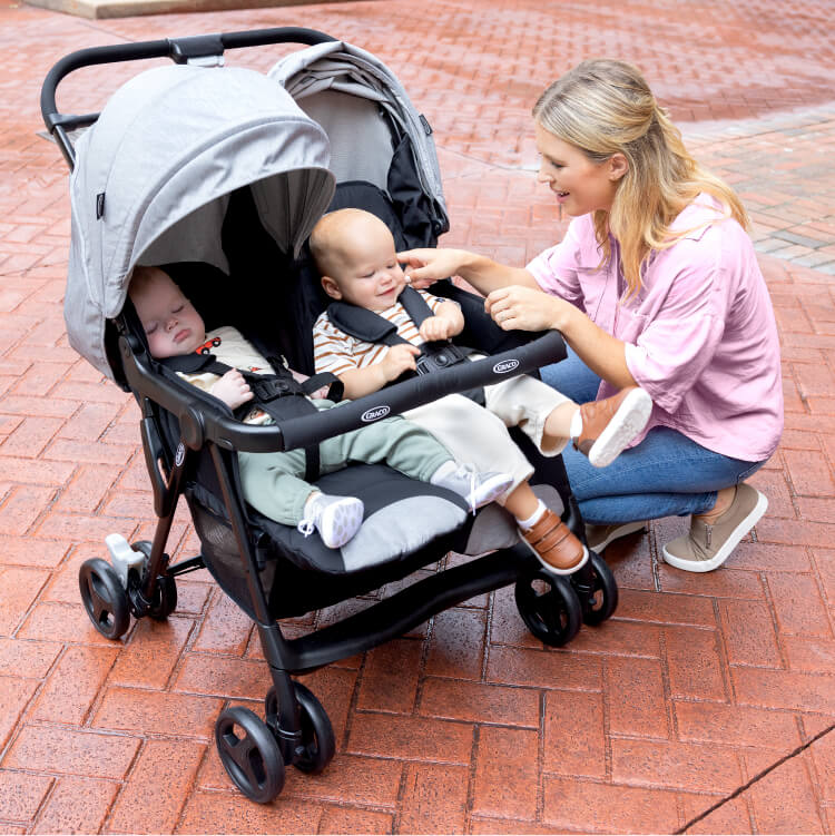 Image of Mum interacting with a smiling baby in Graco DuoRider double stroller while the other baby is asleep with calf support extended and back reclined
