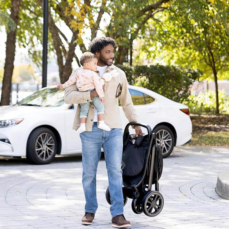 Ein Vater trägt den Graco EeZeFold Buggy mit einer hand, während er mit dem anderen Arm sein Kind hält