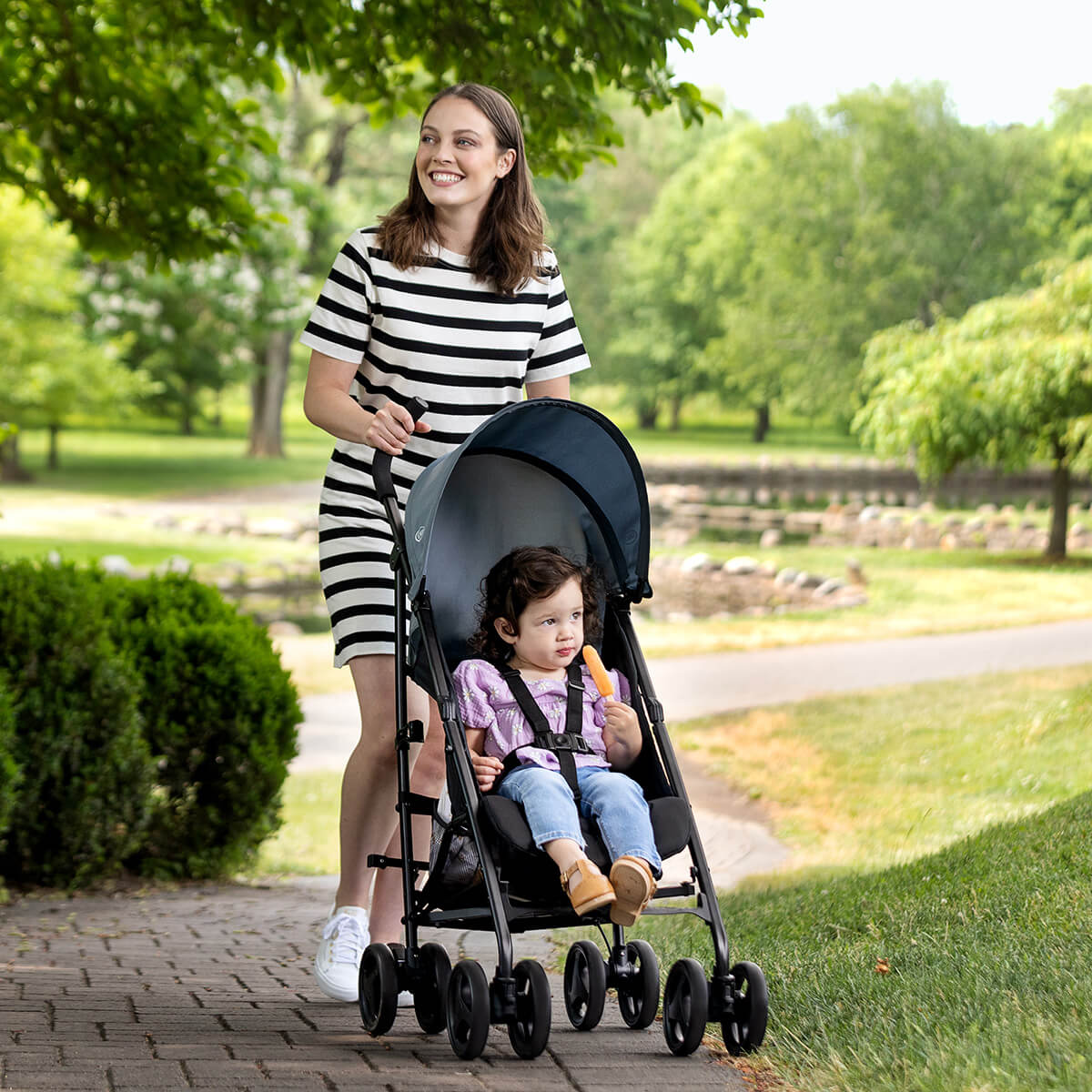 Mum pushing her daughter up a slope in a park in Graco’s lightweight EZLite stroller. 
