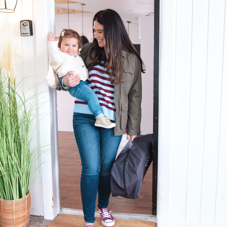 Mutter trägt Kleinkind und das Graco FoldLite™ LX Reisebett mit Bassinet-Einhang durch die Tür.
