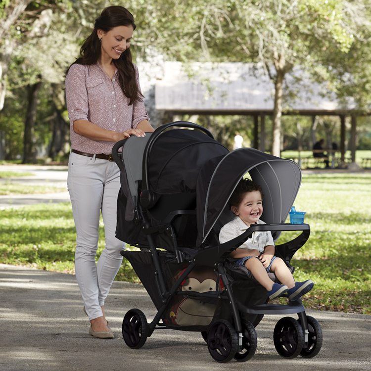 Mum pushing Graco Stadium Duo® with one child in front seat and one child in SnugEssentials infant i-Size car seat in rear seat