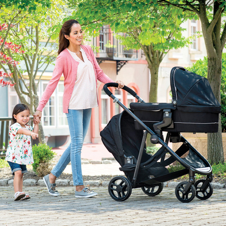 Mum pushing Graco Time2Grow™ double pushchair with Time2Grow™ carrycot attached while walking hand-in-hand with her daughter